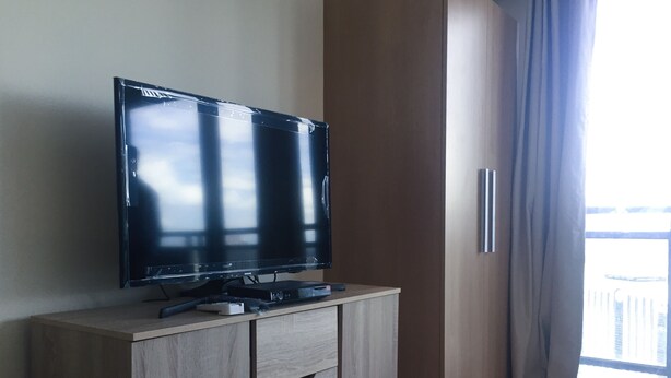 Una habitación moderna con una televisión de pantalla plana sobre un mueble de madera, una mesita de noche pequeña y una ventana con cortinas.
