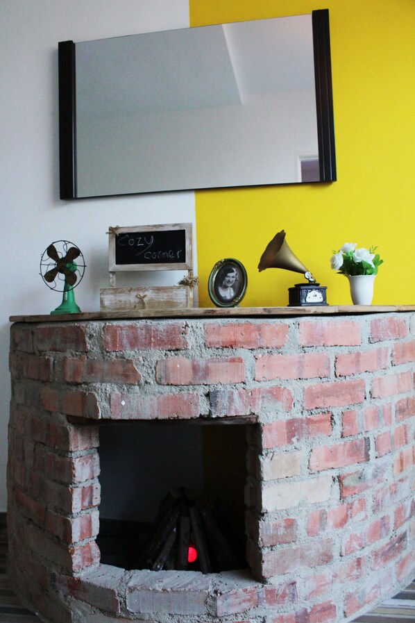 A brick fireplace with a shelf above it holding a fan, a clock, a framed photo, a small chalkboard, and a potted plant.