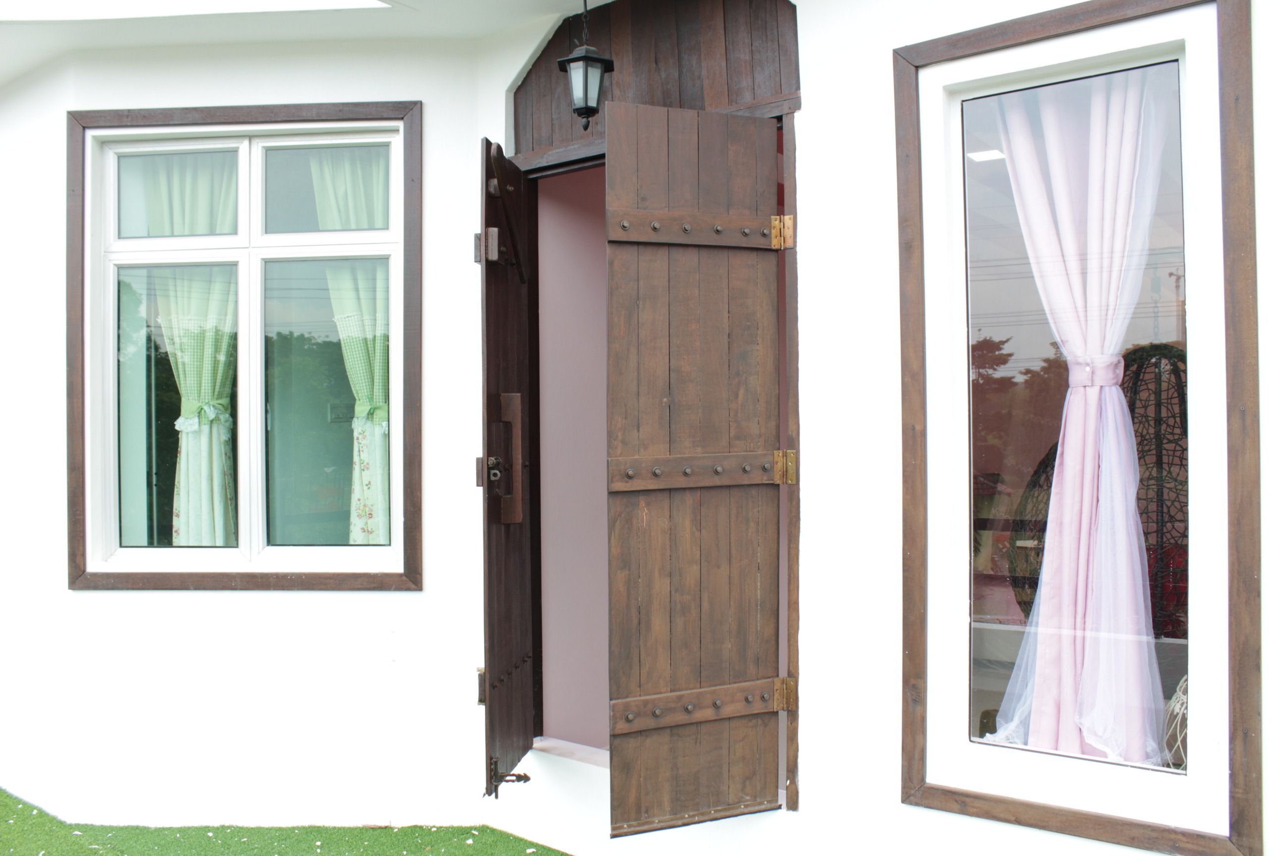 Eine rustikale Holztür mit einem Metallriegel, ein Fenster mit weißem Rahmen und blumigen Vorhängen sowie ein großes Fenster mit einem transparenten Vorhang.