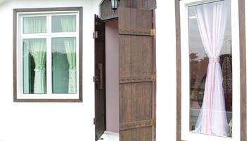 Eine rustikale Holztür mit einem Metallriegel, ein Fenster mit weißem Rahmen und blumigen Vorhängen sowie ein großes Fenster mit einem transparenten Vorhang.