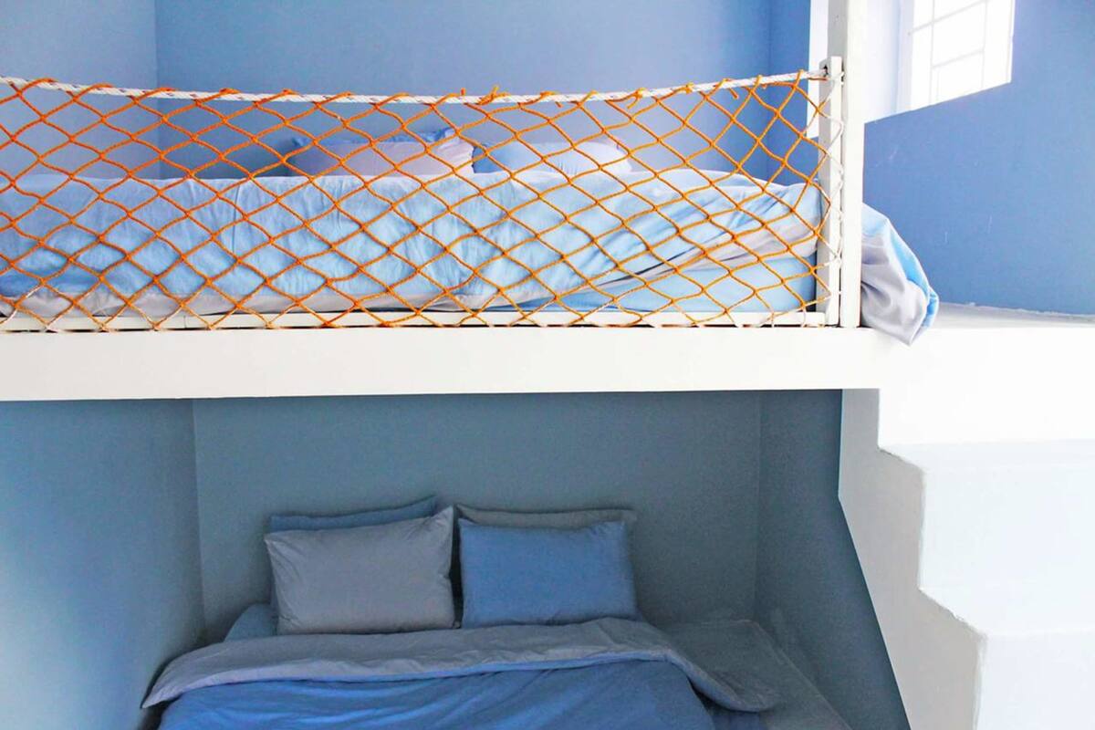 Een stapelbed met een oranje nethek, een onderste bed met blauw beddengoed, en een raam in het bovenste bed.