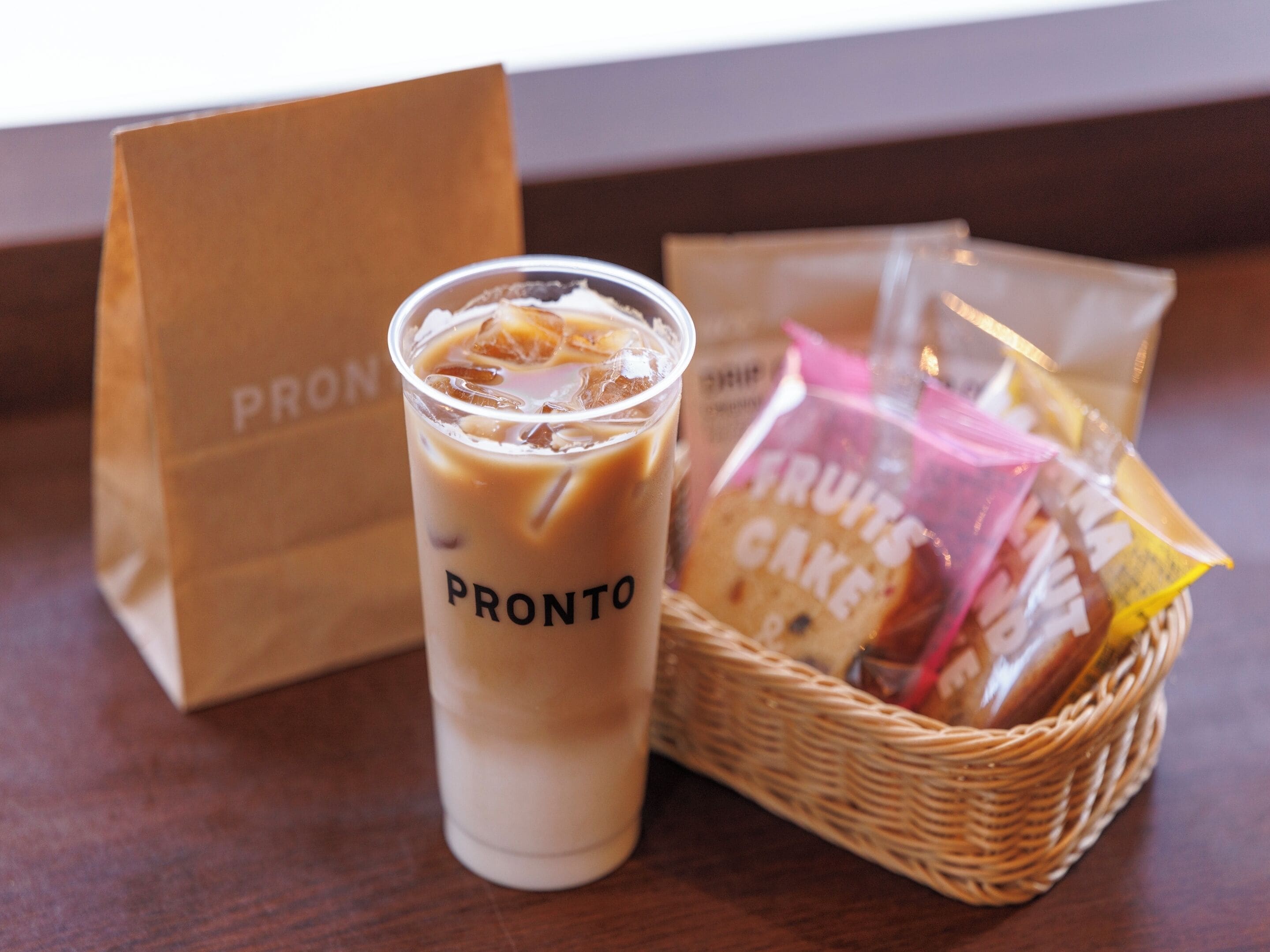 Una bebida en un vaso con el logo de la marca, una bolsa de papel marrón y una cesta con productos envasados.