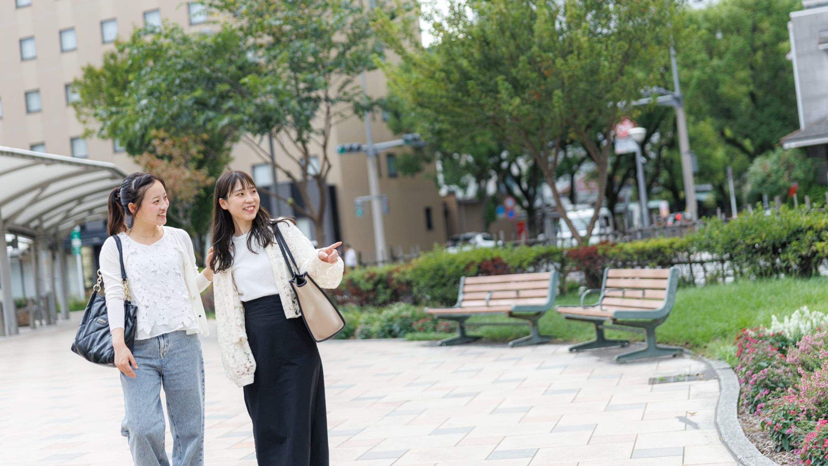 Two individuals walking on a paved pathway with benches and greenery in the background.