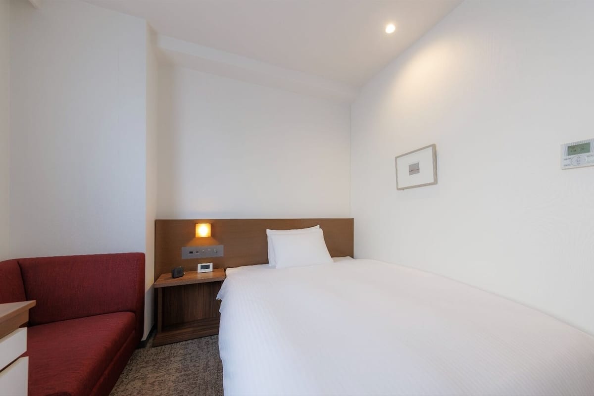 A hotel room with a bed, a red sofa, a nightstand with a lamp, and a wall-mounted thermostat.
