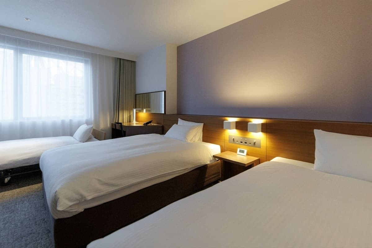 A hotel room with two beds, a bedside table with a lamp, a mirror, and a window with sheer curtains.