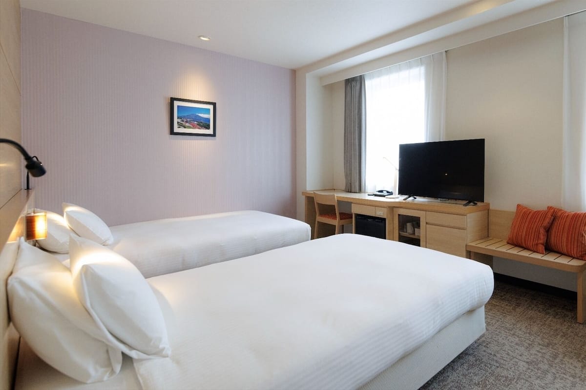 A hotel room with two beds, a desk with a TV, a small table, and a window with curtains.