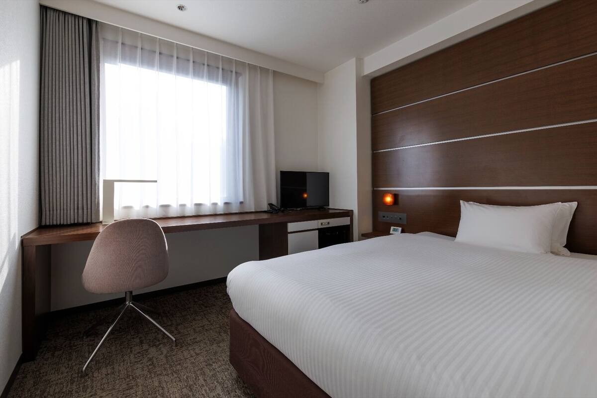 A hotel room with a bed, a desk with a computer, a chair, and a window with curtains.