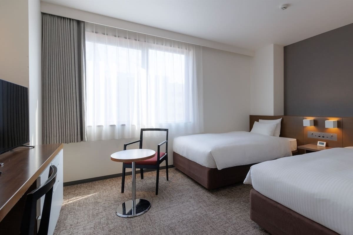 A hotel room with two beds, a desk, a chair, and a TV.