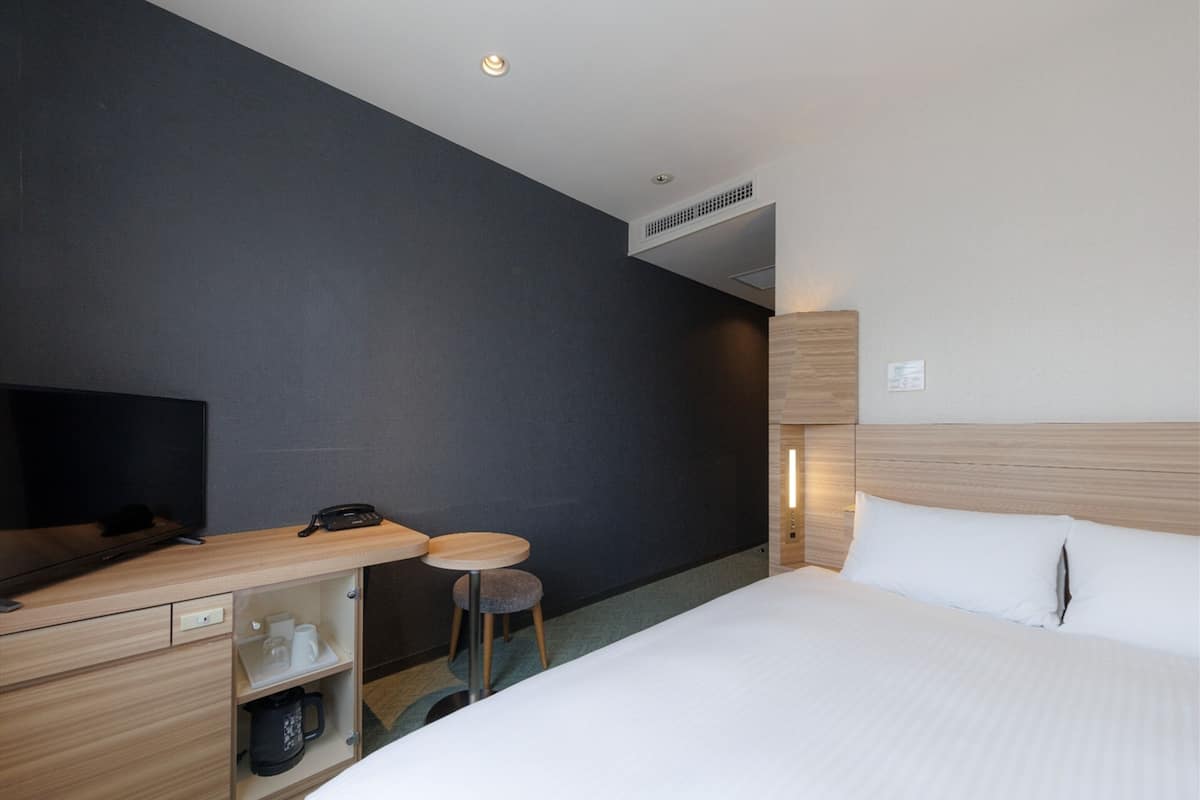 A hotel room with a bed, a desk with a telephone, a television, and a small stool.