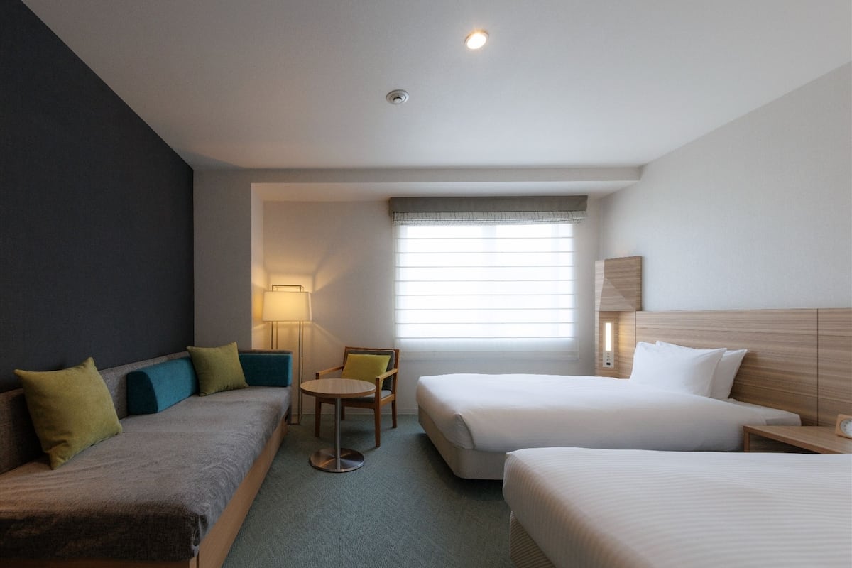 A hotel room with two beds, a sofa, a chair, a lamp, and a window with blinds.