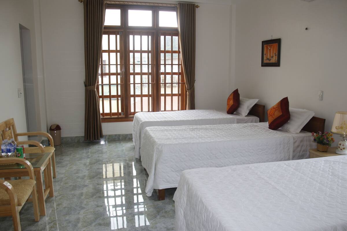A hotel room with two beds, a dining table with chairs, a window with curtains, and a wall-mounted picture.