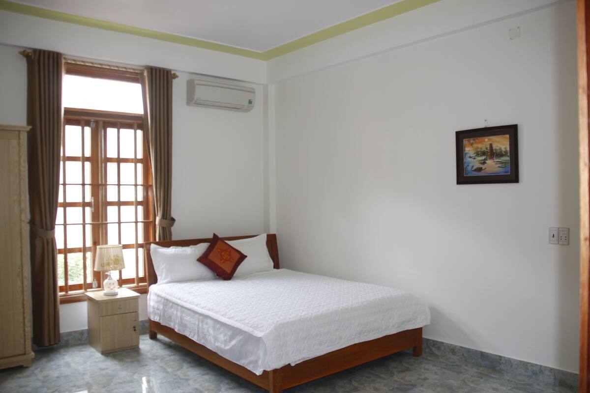 A bedroom with a wooden bed, a nightstand, a window with curtains, and a wall-mounted air conditioner.
