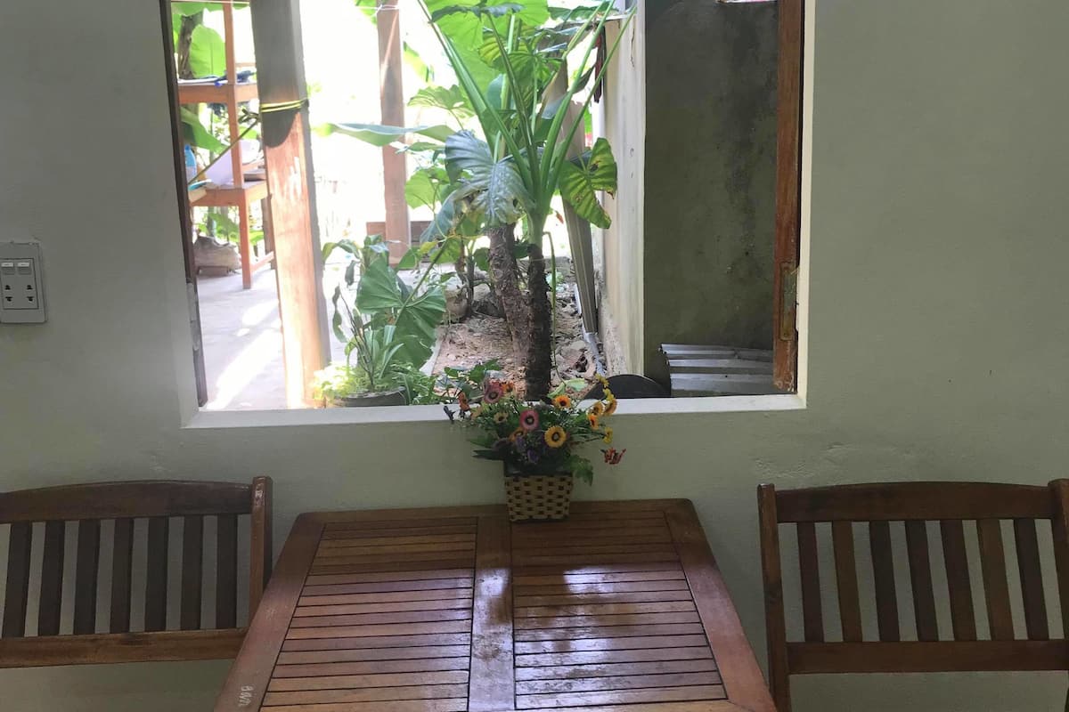 A wooden table with two chairs, a window with a view of a wooden structure and plants outside, and a small flower arrangement on the table.
