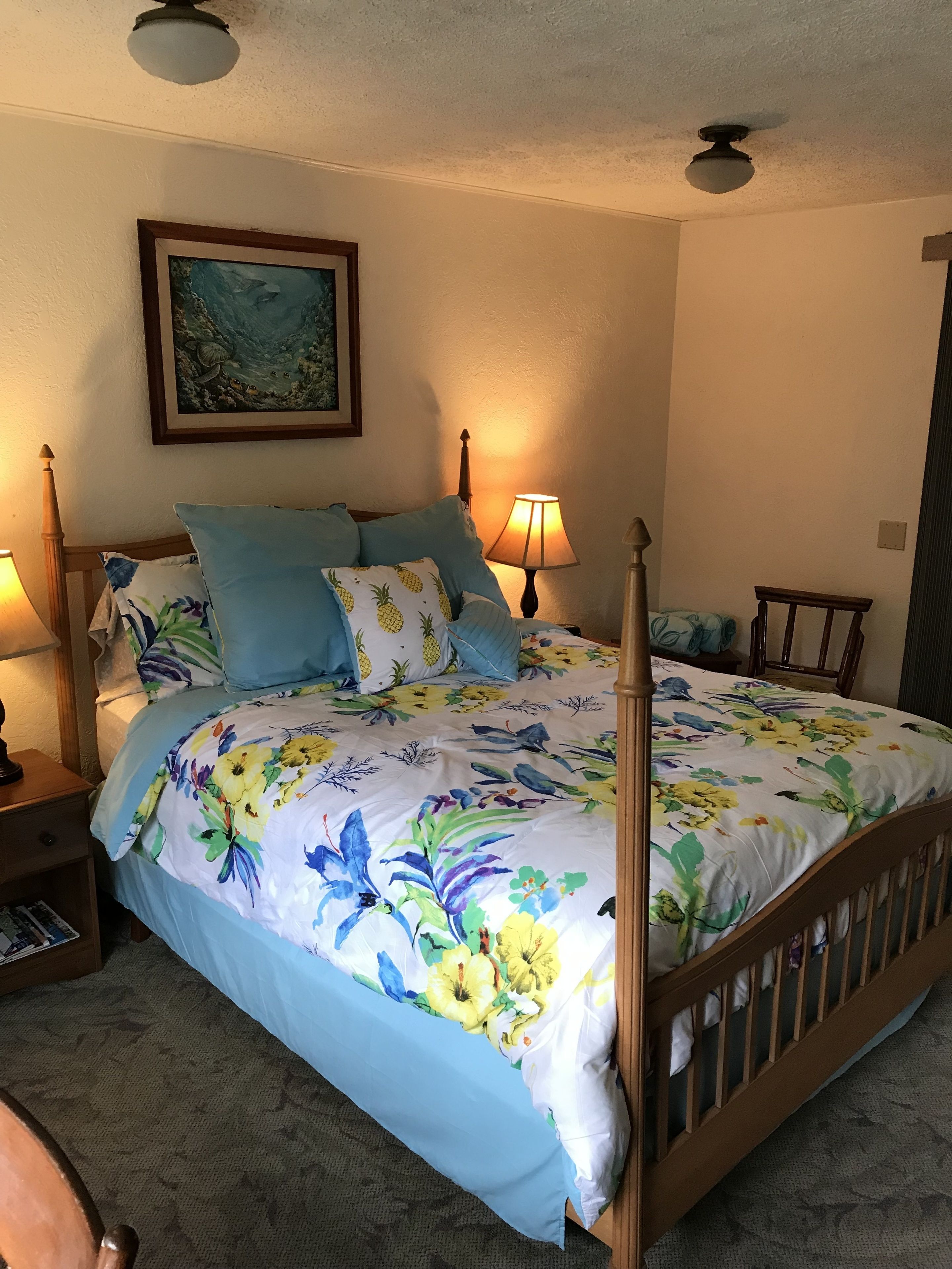 Ein Schlafzimmer mit blumigem Bettbezug, einem Holz-Kopfteil, zwei Nachttischlampen, einem gerahmten Bild an der Wand und einem Deckenventilator.