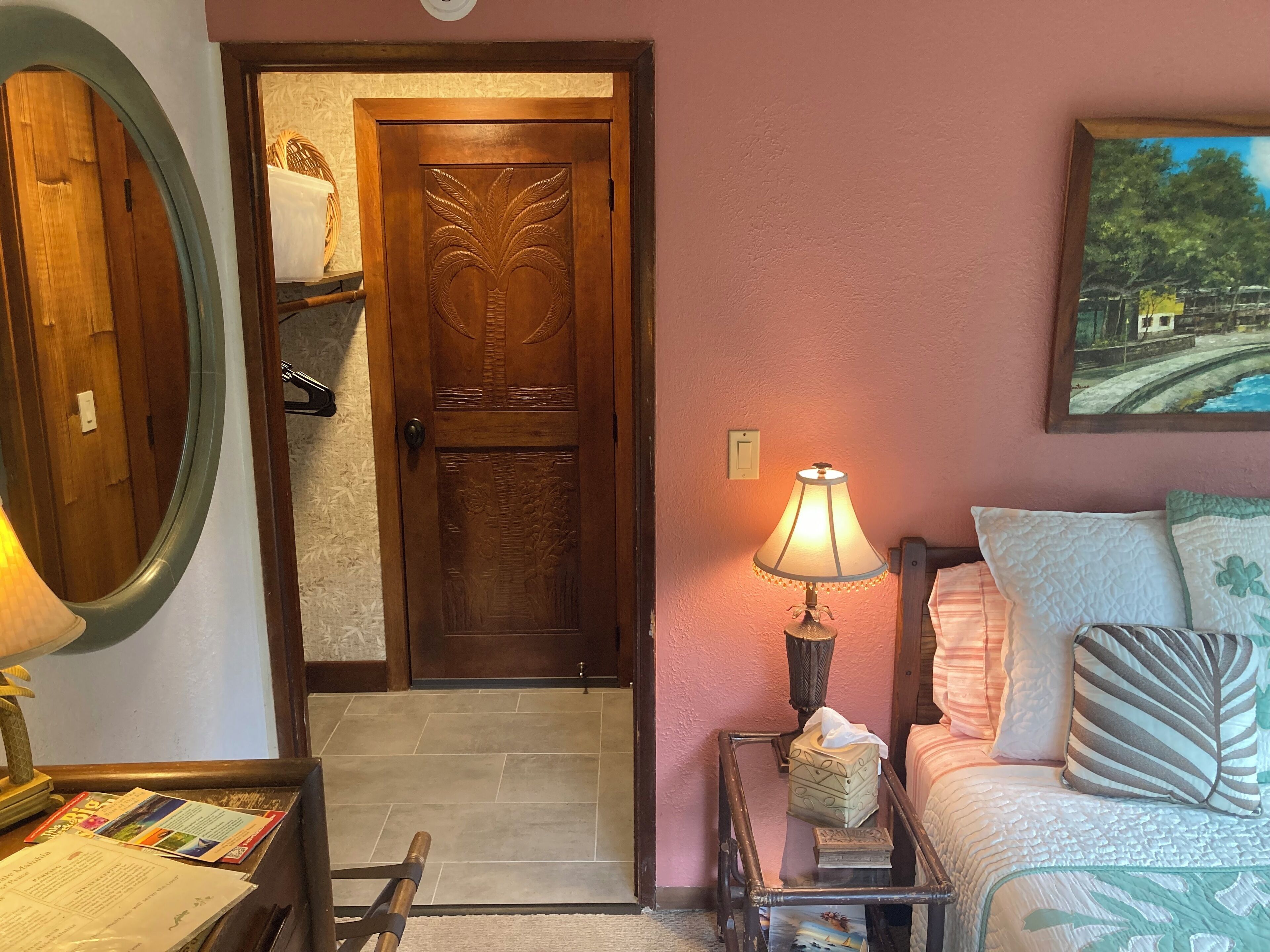 A cozy room with a wooden door, a bed with patterned pillows, a bedside table with a lamp, and a framed painting on the wall.