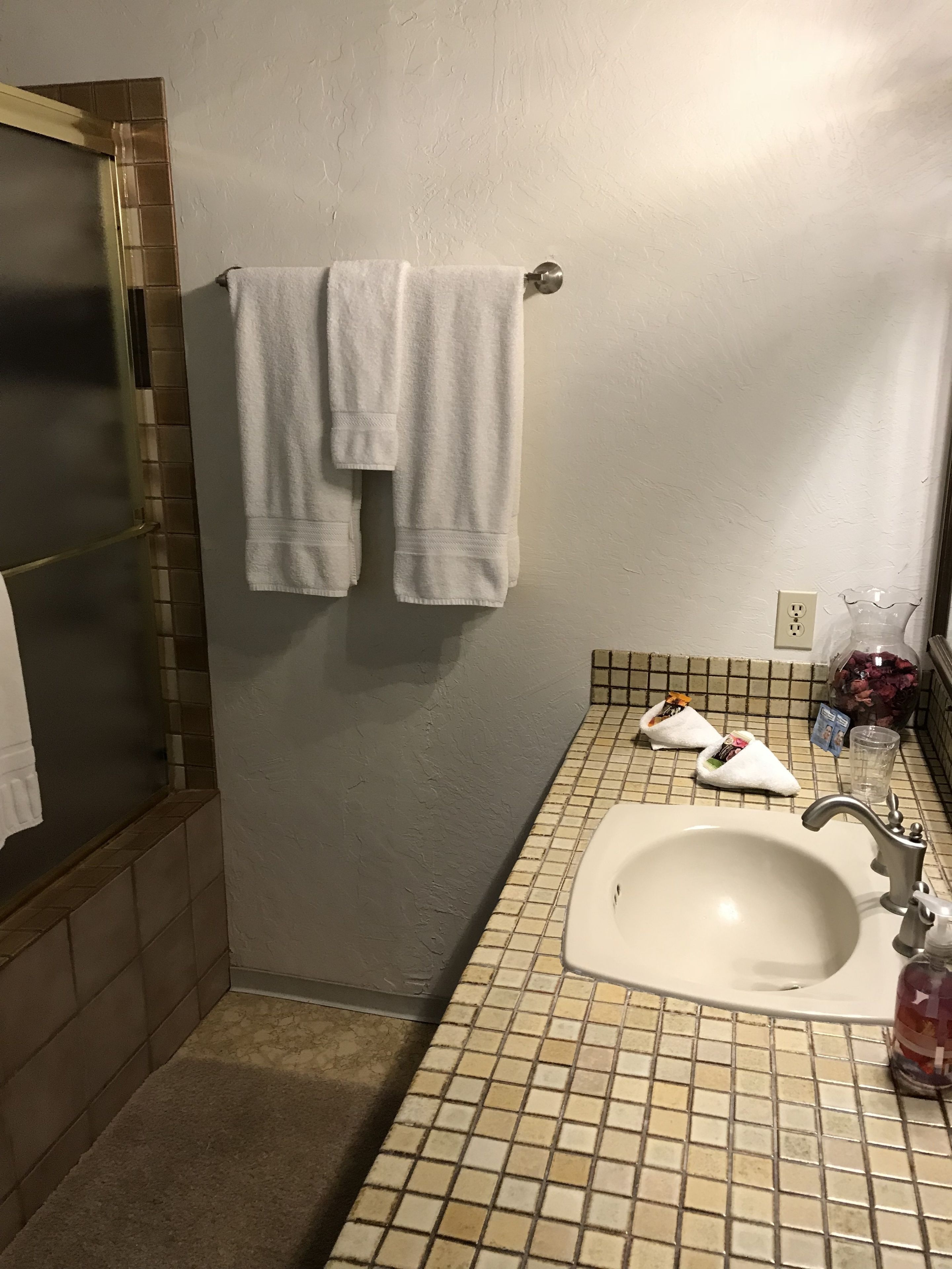 Une salle de bain avec un lavabo carrelé, un porte-serviettes avec deux serviettes, un verre d’eau et des articles de toilette posés sur le comptoir.