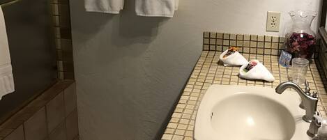 Une salle de bain avec un lavabo carrelé, un porte-serviettes avec deux serviettes, un verre d’eau et des articles de toilette posés sur le comptoir.