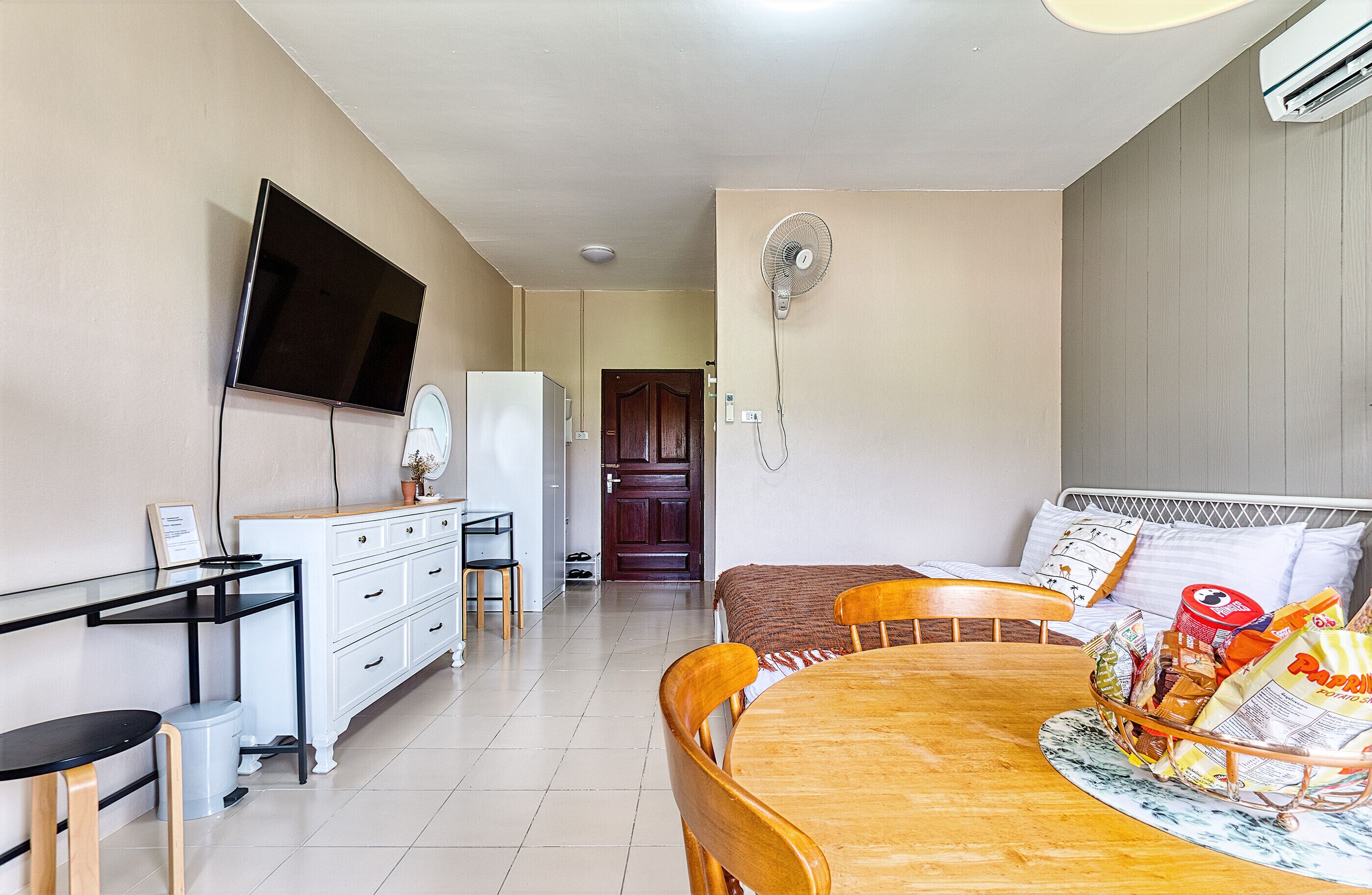 Una stanza con TV a muro, un frigorifero, un tavolo da pranzo con sedie, una ventola e un letto con cuscini.