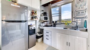 Cocina compacta con refrigerador de acero inoxidable, microondas y cafetera. Hay estantes con frascos y un bote de basura. Una ventana ofrece vistas al exterior.