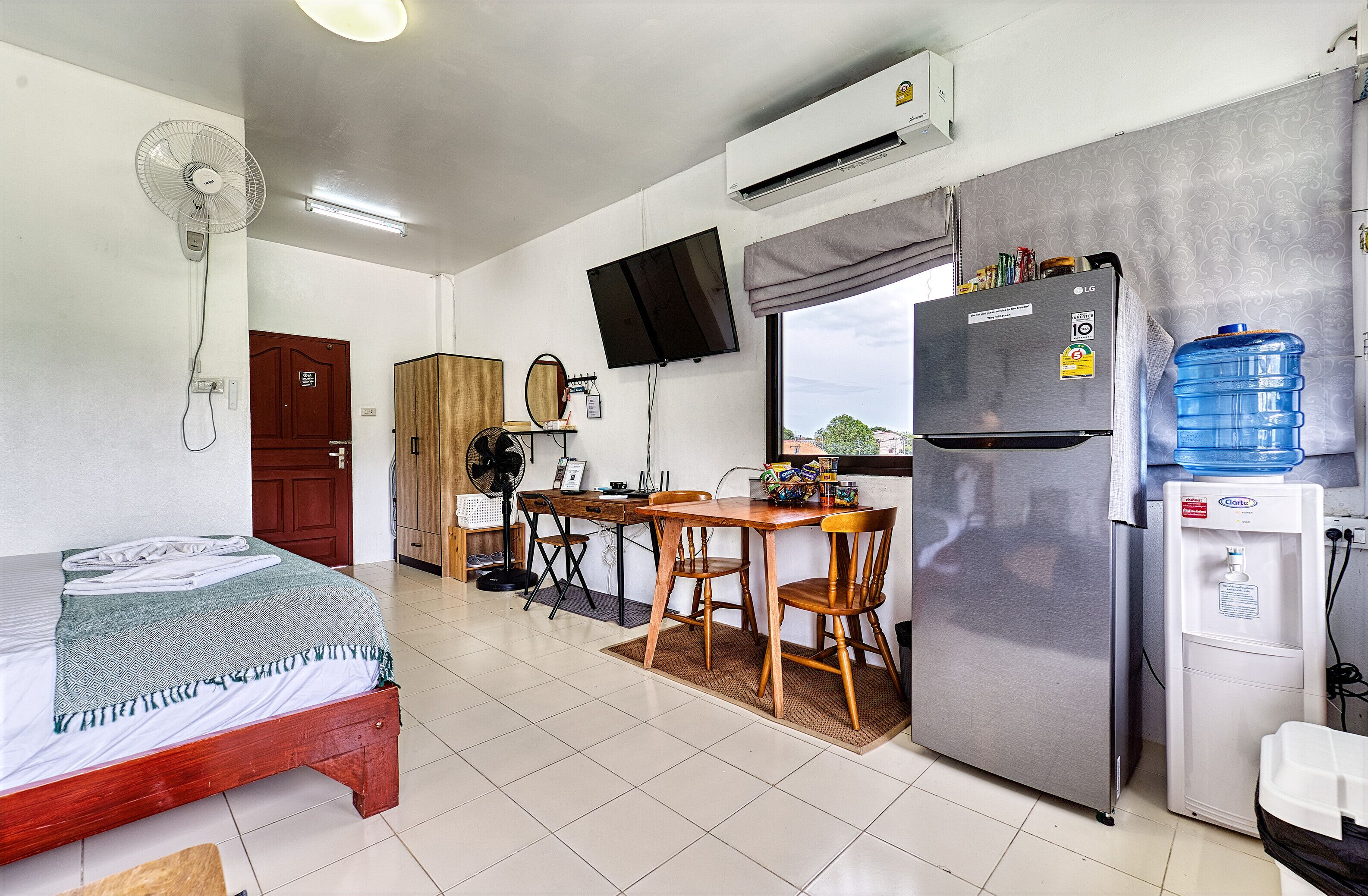 A room with a bed, a desk with a chair, a refrigerator, a water dispenser, a TV, and a fan.