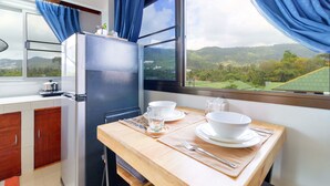 Una cocina con refrigerador, una mesa de comedor dispuesta con vajilla blanca y un vaso, y vista a colinas verdes a través de una ventana con cortinas azules.