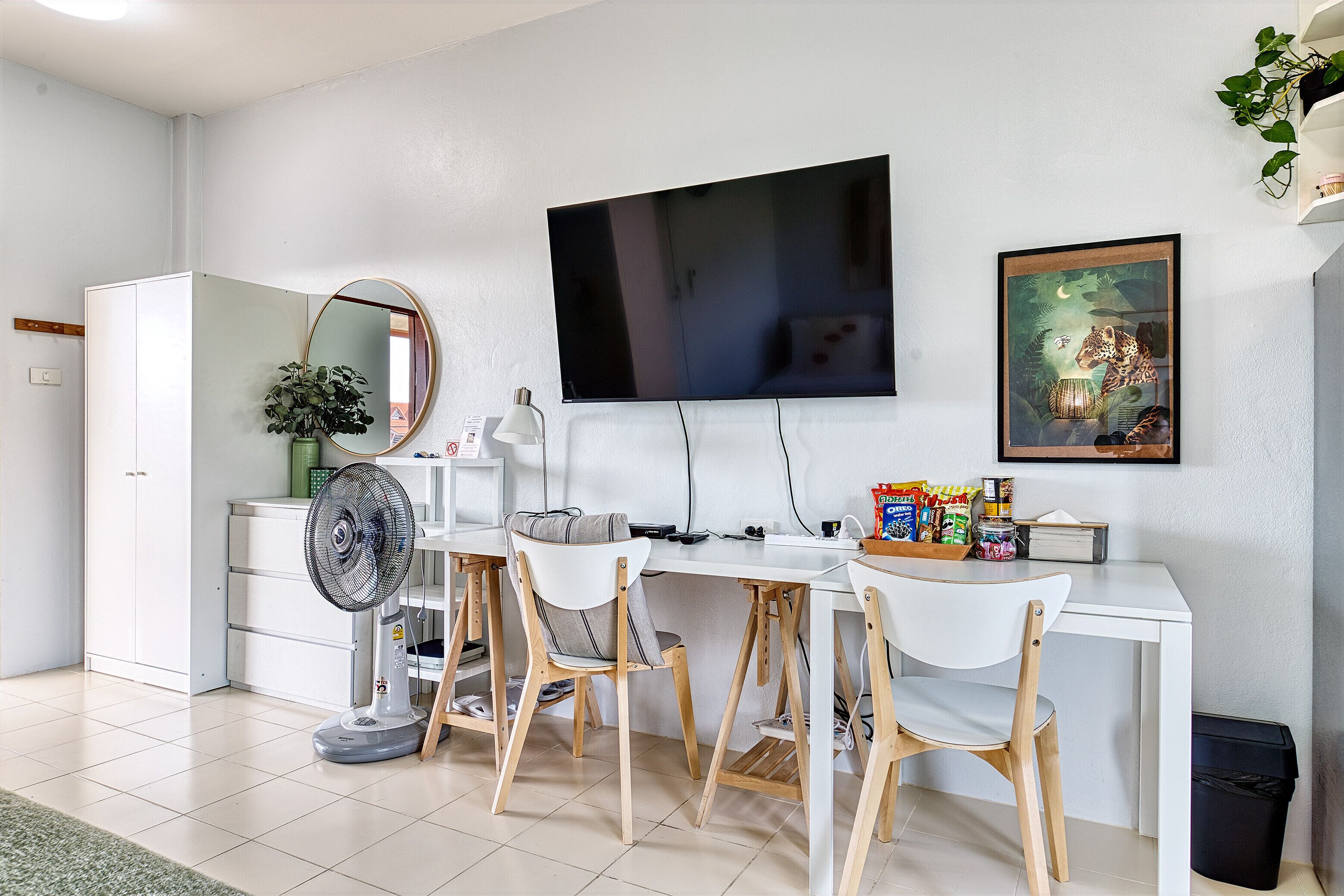 A modern, well-lit room with a white desk, two chairs, a large flat-screen TV, a mirror, and a framed picture on the wall.