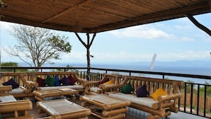 A covered outdoor seating area with bamboo furniture and colorful cushions, overlooking a scenic ocean view. - Bataran Garden Cottage (Penida Island)