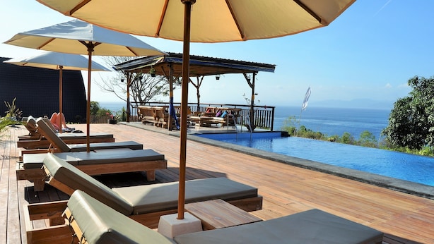 A wooden deck with lounge chairs and umbrellas overlooking a swimming pool and ocean view. - Bataran Garden Cottage (Penida Island)