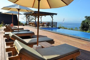 Une terrasse en bois avec des chaises longues et des parasols, offrant une vue sur une piscine et l’océan.