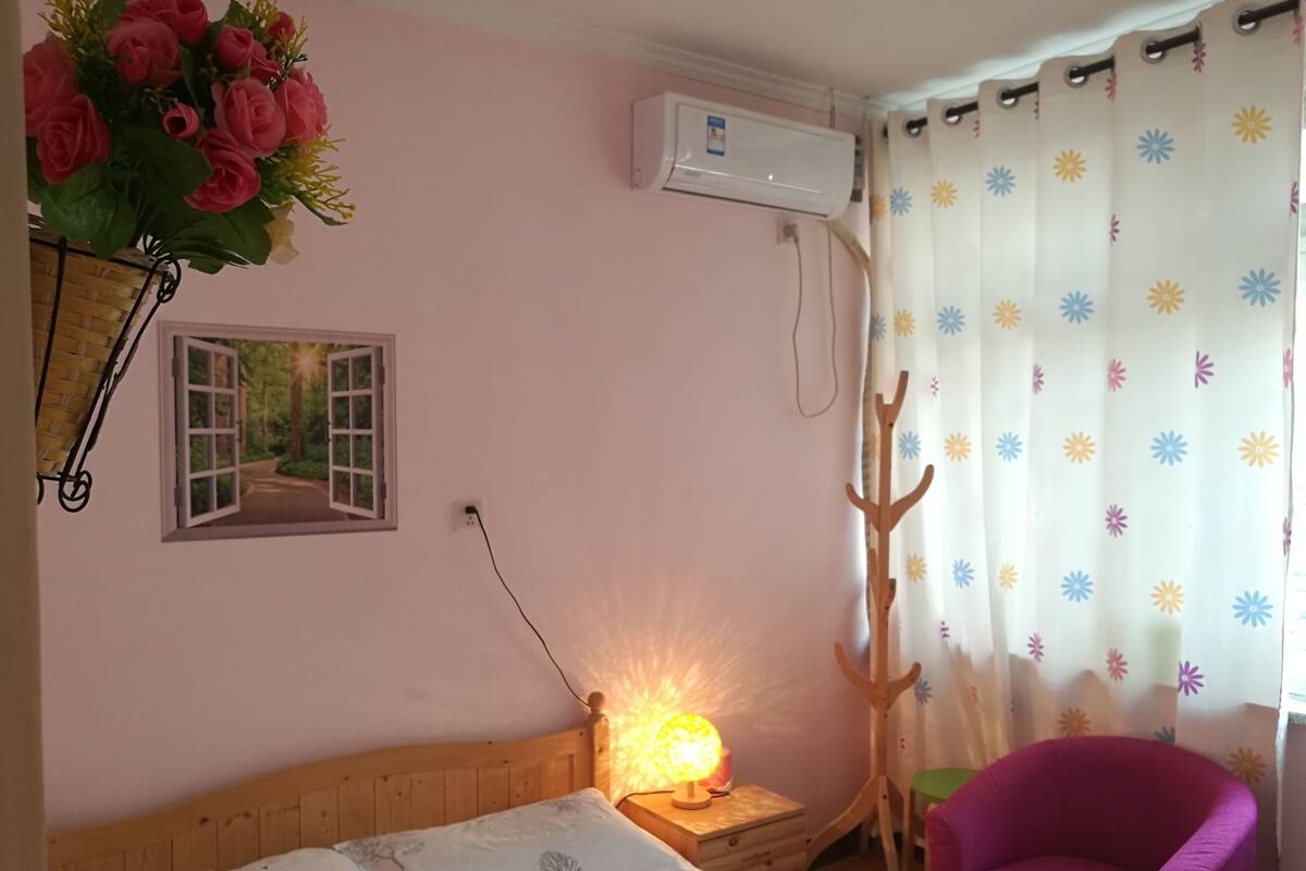 Un dormitorio con cama, mesita de noche con lámpara, una silla, aire acondicionado de pared y ventana con vista a árboles.