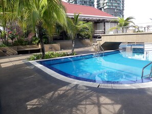 A swimming pool with a tiled edge, surrounded by palm trees and a building with a red roof in the background.