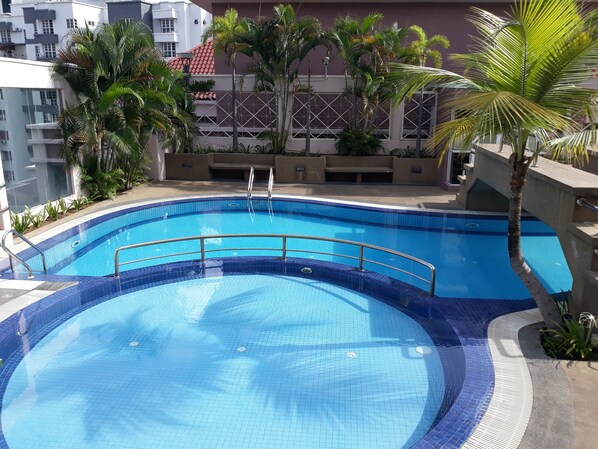 A circular swimming pool with a tiled edge, surrounded by palm trees and a building with a red-tiled roof in the background.