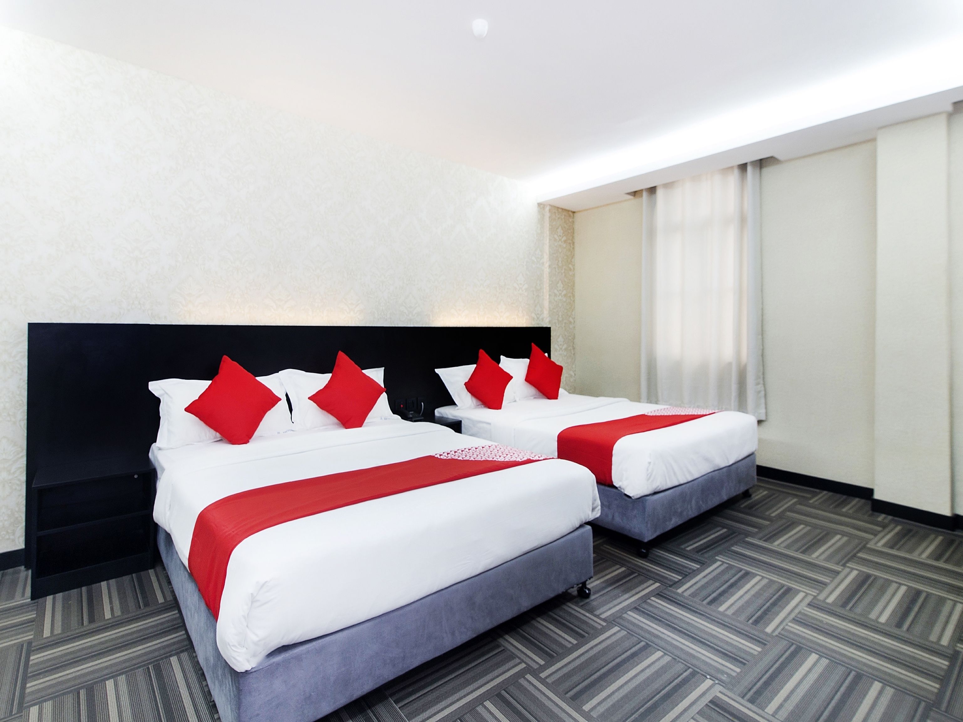 Habitación de hotel con dos camas, cada una con ropa blanca y almohadas rojas, un cabecero negro y una alfombra gris.