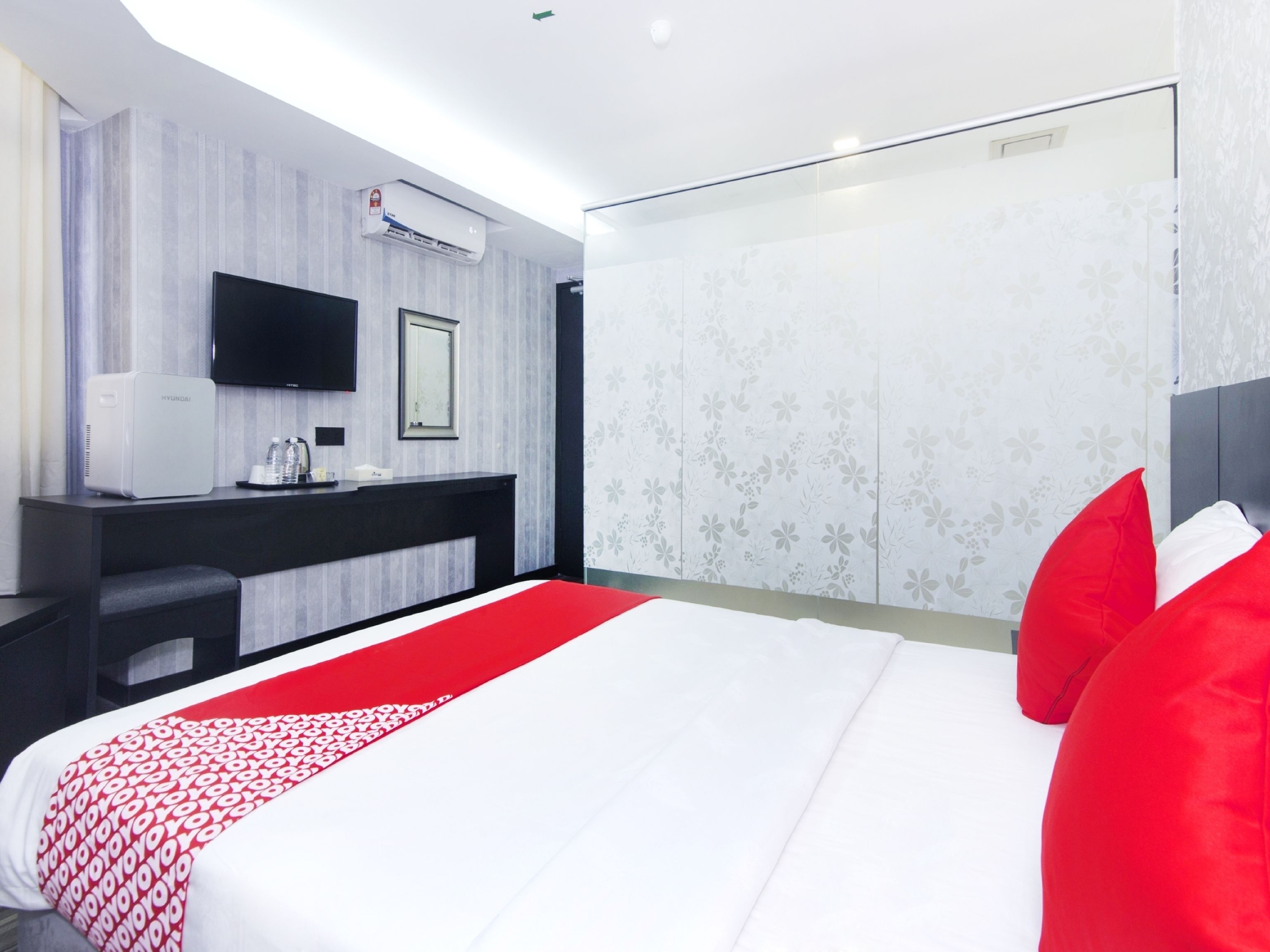 A modern hotel room with a large bed, a flat-screen TV, a minibar, and a large window.