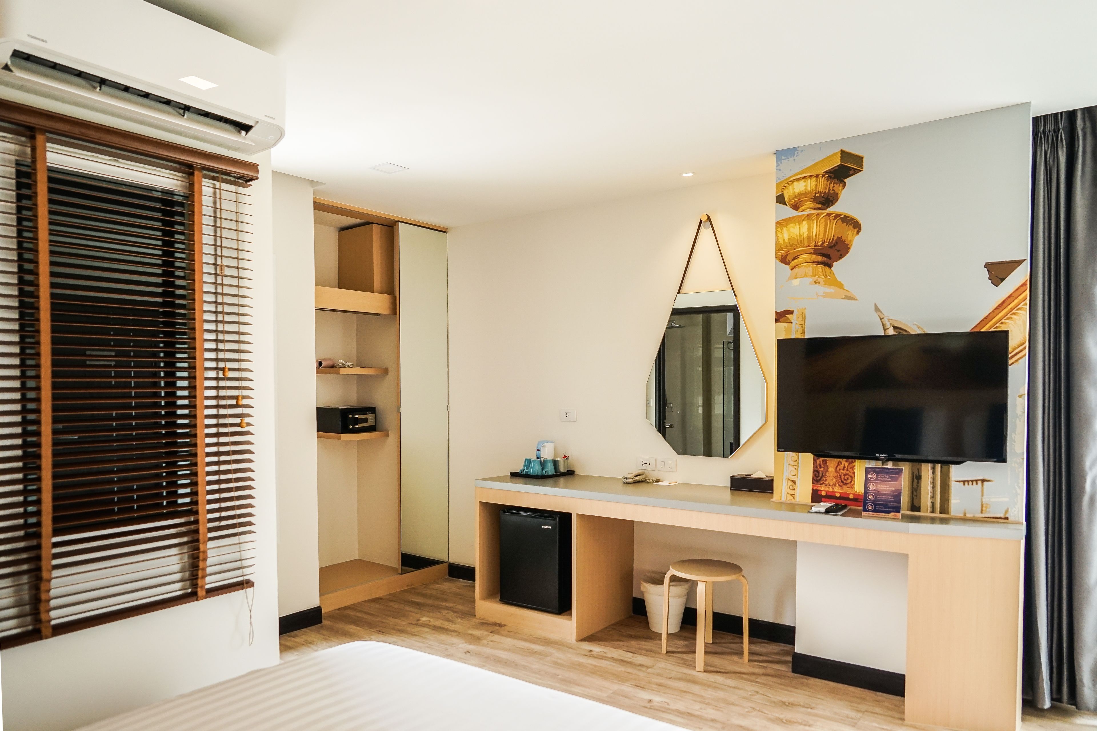 Ein modernes Hotelzimmer mit Schreibtisch aus Holz, einem Fernseher, einem großen Spiegel und einem Fenster mit Jalousien.