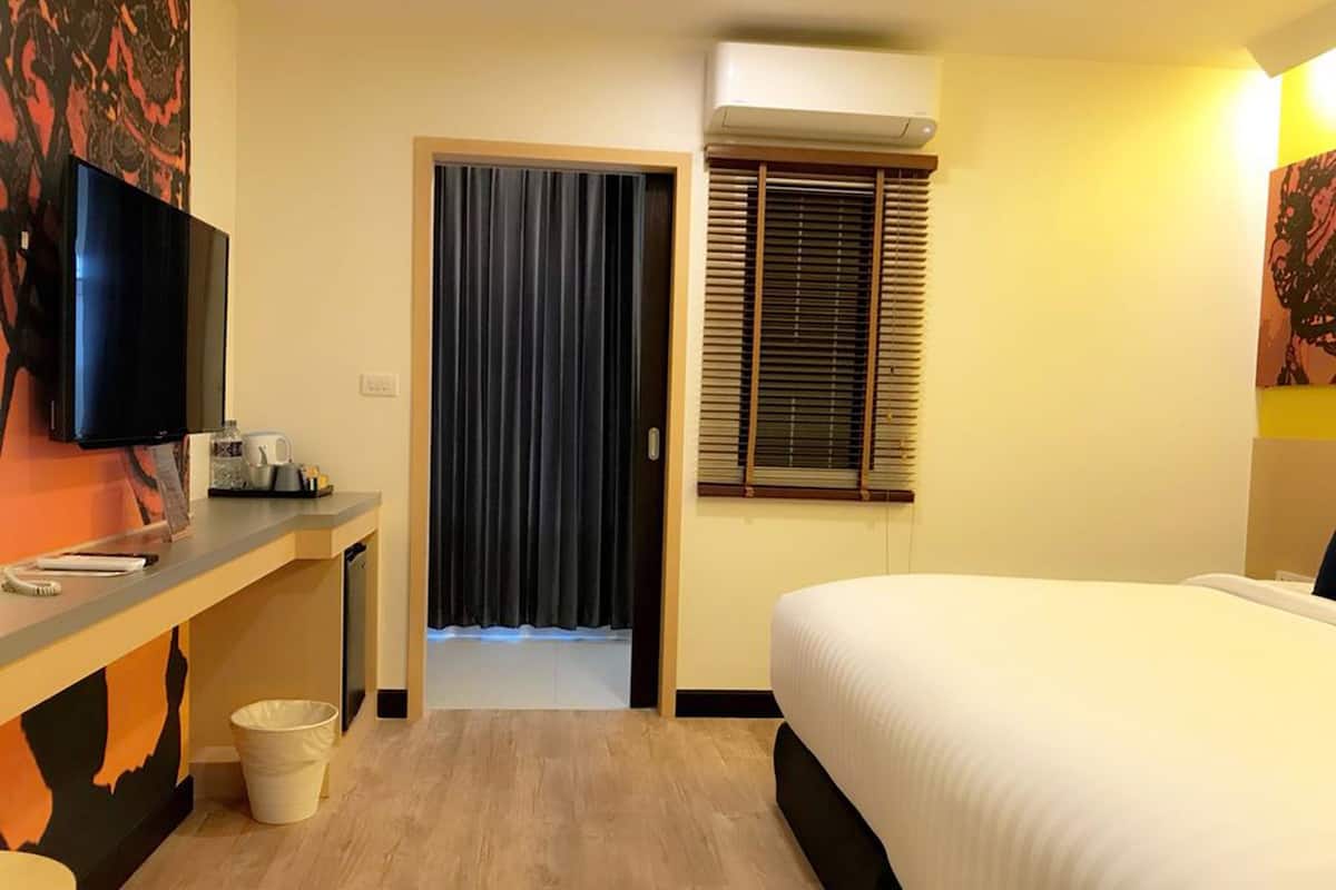 A hotel room with a bed, a desk with a flat-screen TV, a window with blinds, and an air conditioning unit.