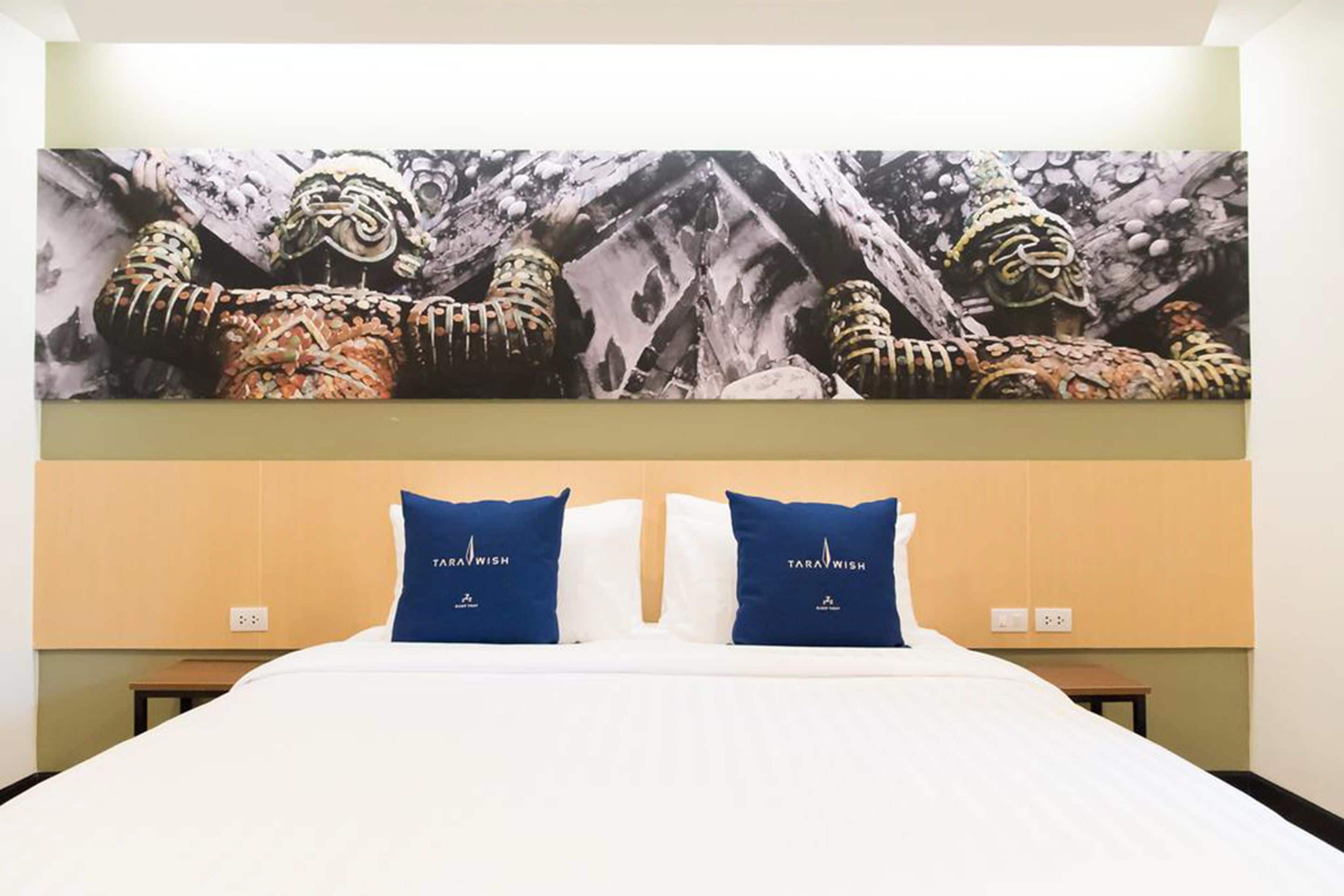 A hotel room with a large bed, two blue pillows with 'TARAWISH' branding, and a wall mural depicting a traditional scene.