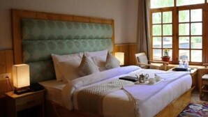 A bedroom with a large bed, wooden headboard, bedside tables with lamps, a small table with a plant, and a window with curtains. - Himalayan Retreat Karzoo (Leh)