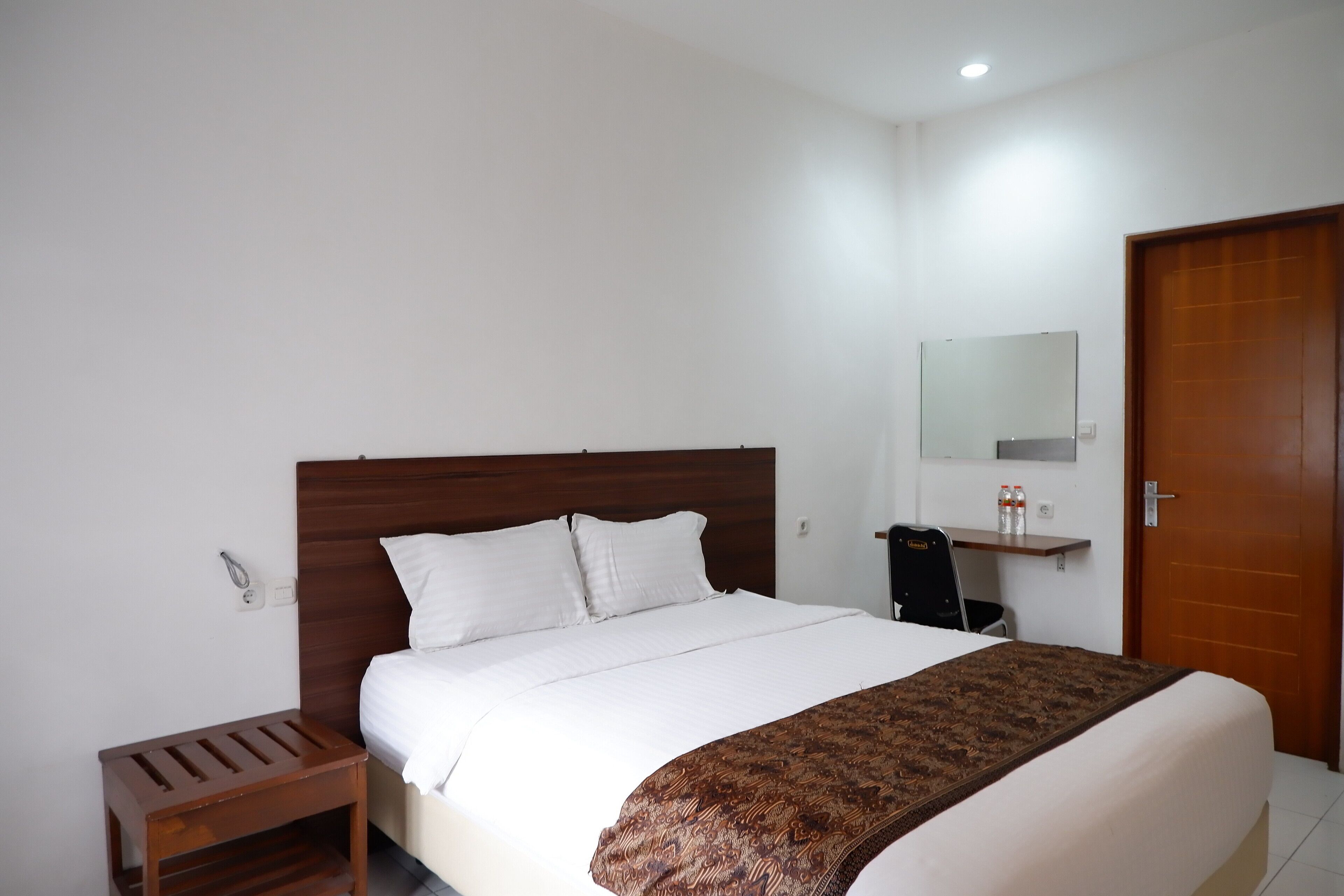 A neatly made bed with white bedding and a patterned throw, a wooden nightstand, a small desk with a chair, a mirror on the wall, and a wooden door.