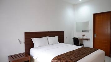 A neatly made bed with white bedding and a patterned throw, a wooden nightstand, a small desk with a chair, a mirror on the wall, and a wooden door.