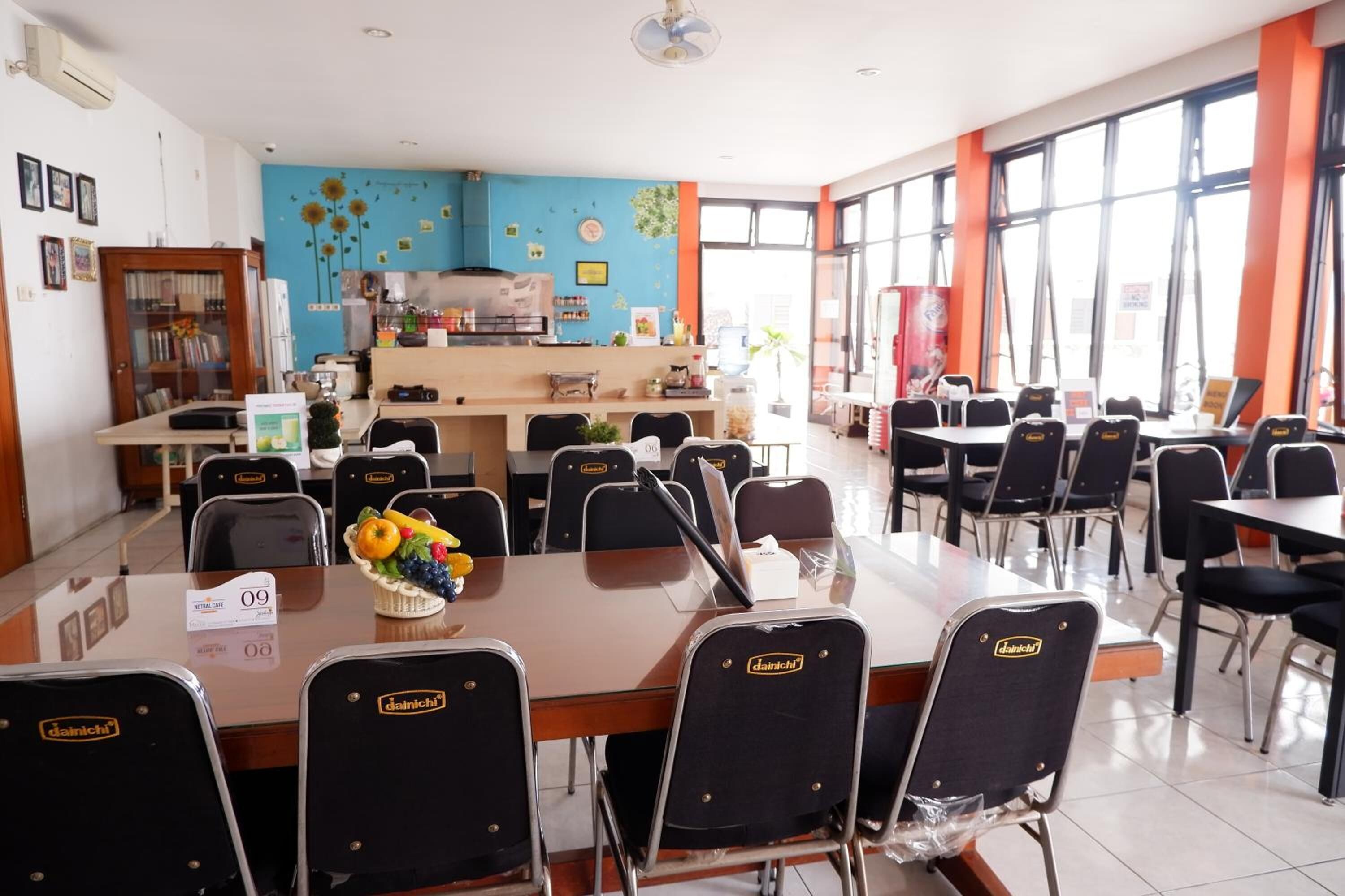 A dining area with tables and chairs, a kitchen in the background, and large windows.