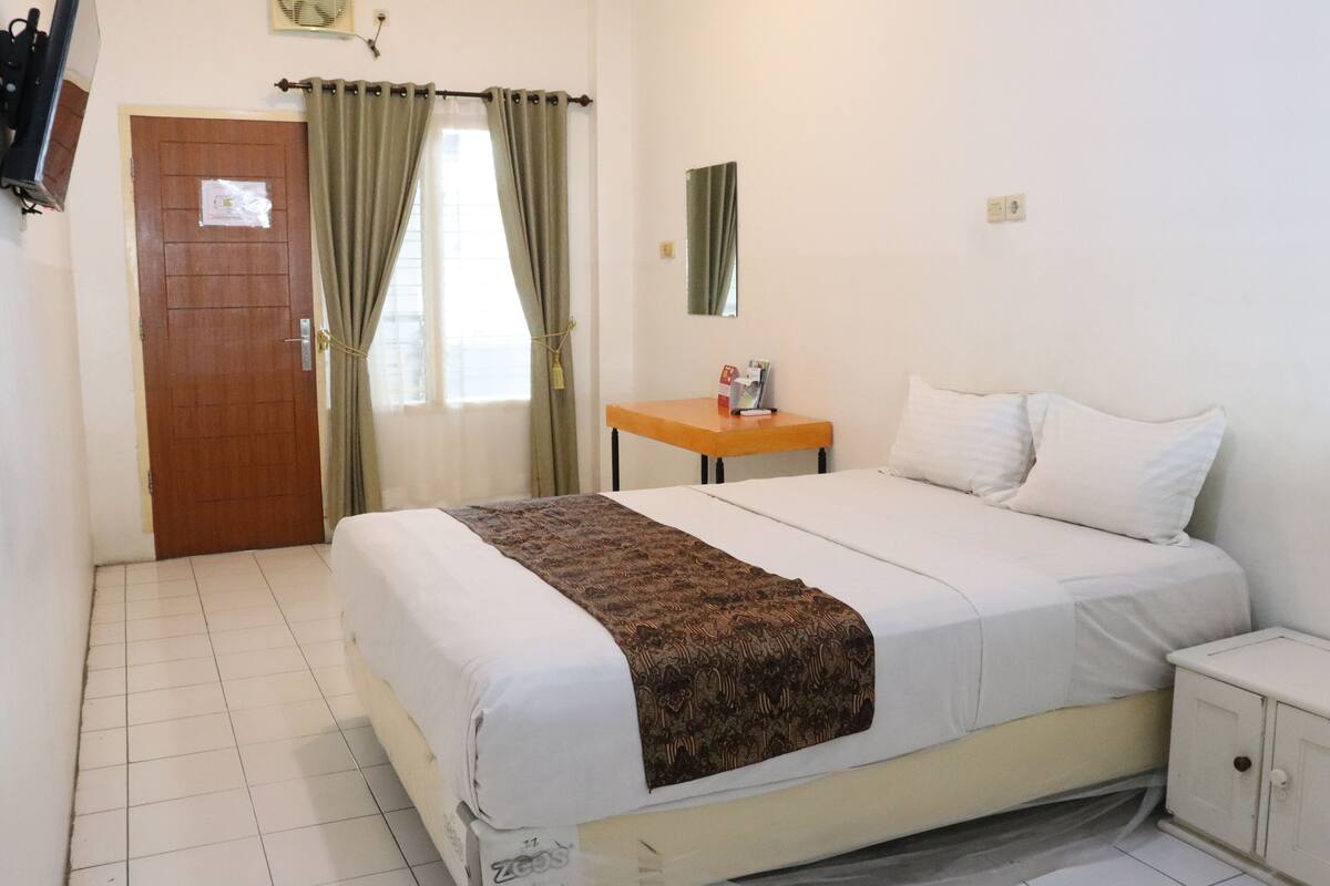 Um quarto com cama, mesa de cabeceira, espelho, porta e janela com cortinas.