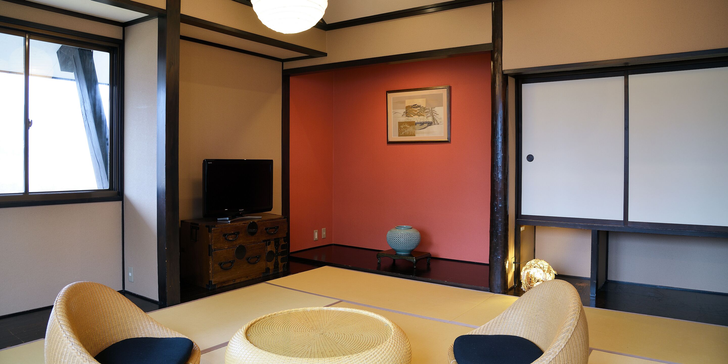 A traditional Japanese room with tatami flooring, a television on a wooden cabinet, a red accent wall, and a shoji screen.