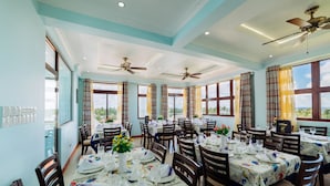 A dining area with multiple tables set for service, ceiling fans, and large windows offering a view. - Rekidhiye Inn (Hithadhoo)