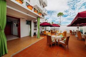 An outdoor dining area with tables and chairs, red umbrellas, and potted plants on the balcony.