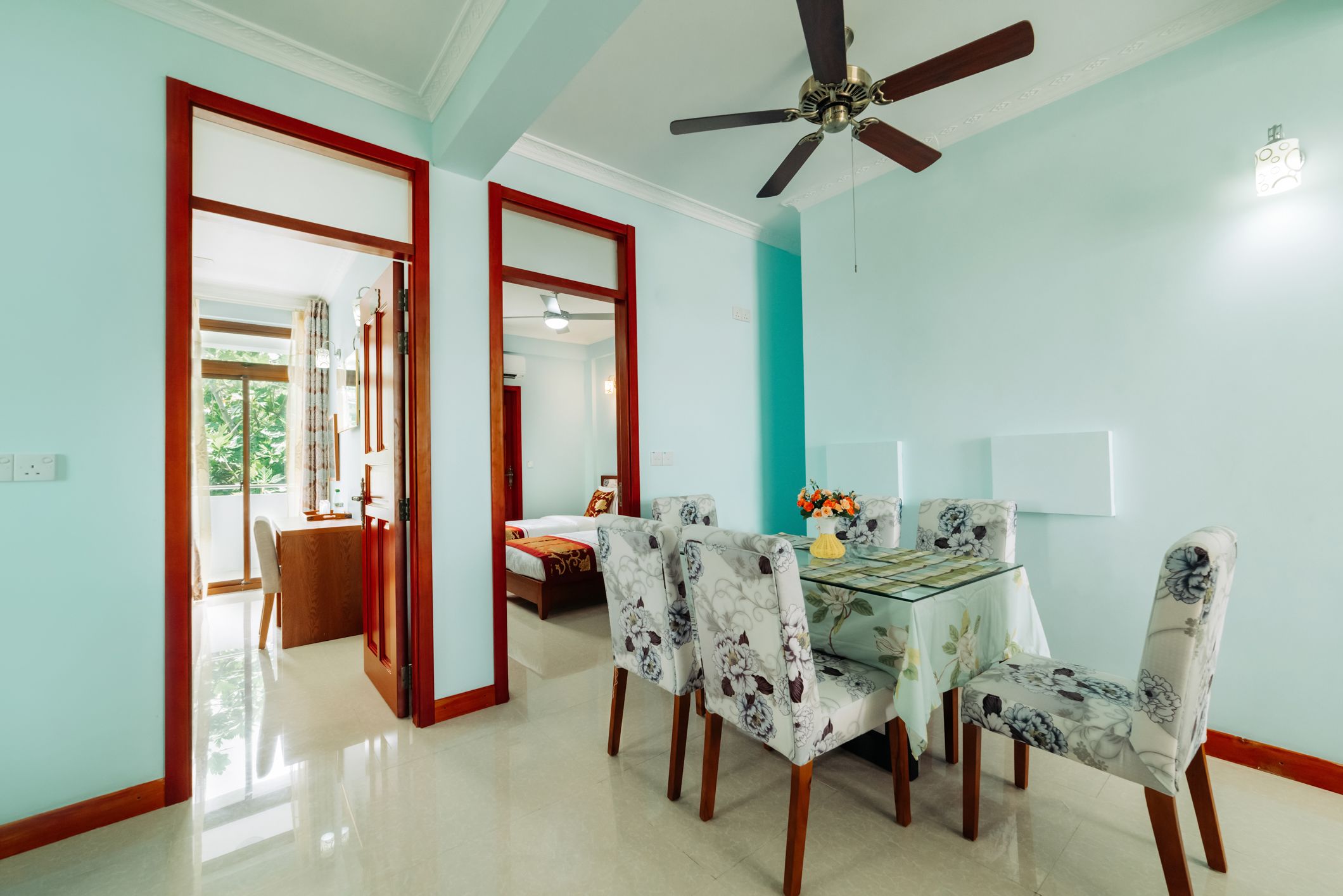 A dining area with a glass table, floral chairs, a ceiling fan, and a view of a bedroom through an open doorway.
