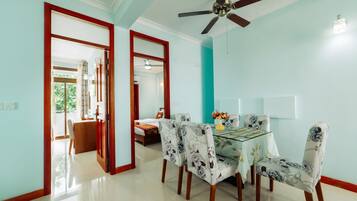 A dining area with a glass table, floral chairs, a ceiling fan, and a view of a bedroom through an open doorway.