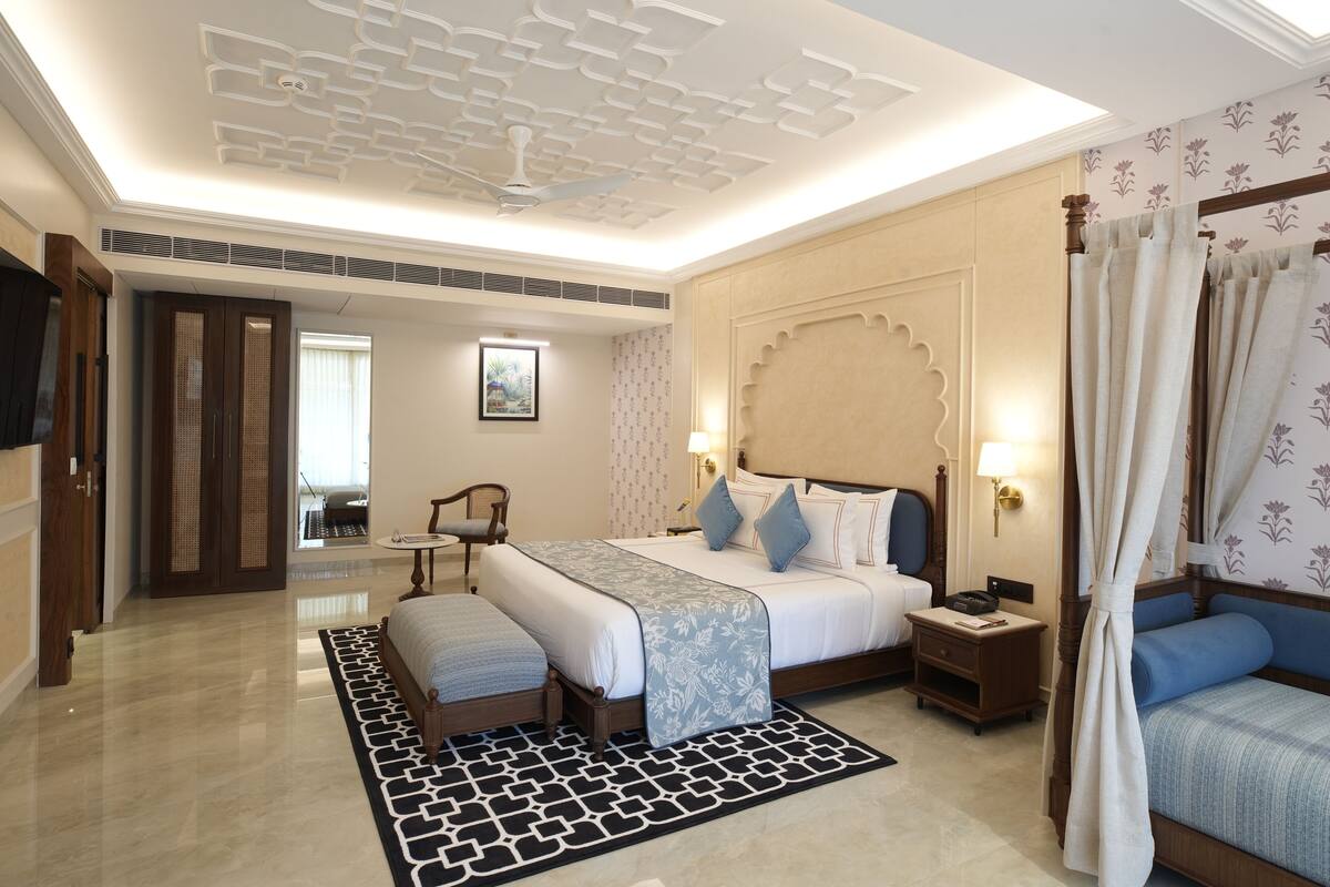 A hotel room with a large bed, a bedside table, a nightstand, a chair, a desk, a window, a ceiling fan, and a bathroom visible through an open door.