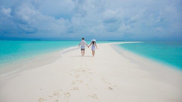 Una pareja caminando sobre una playa arenosa, con agua turquesa cristalina y un cielo nublado.