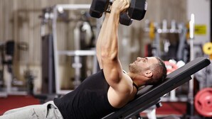 Una persona recostada en un banco dentro de un gimnasio, levantando una pesa.